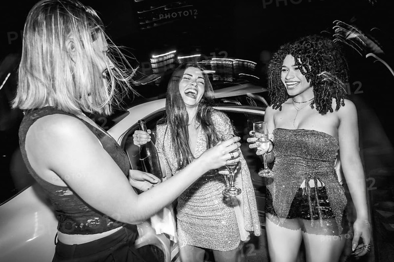 Group of fashionable young women laughing and toasting with champagne in a chic outdoor nightlife setting, enjoying a spontaneous, joyful evening of friendship and celebration