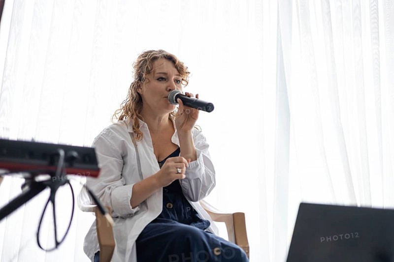 A young Caucasian woman with curly hair sings into a microphone while seated in a cozy home setting. Soft light filters through sheer curtains, creating a warm atmosphere