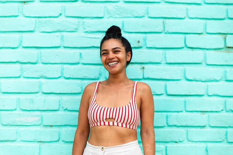 A joyful young Latina woman smiling brightly in a vibrant striped crop top, standing against a turquoise brick wall in Mancora, Peru, radiating summer vibes