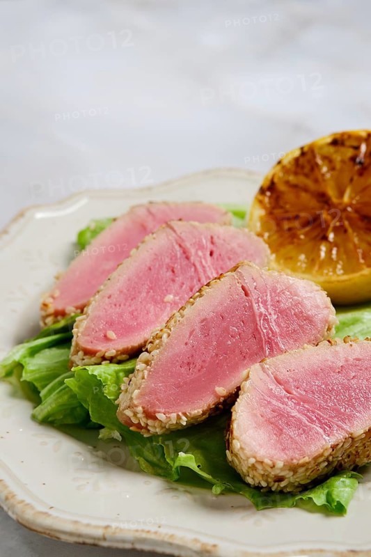 Sliced seared tuna rests on bed of lettuce with sesame crust. Grilled lemon half sits beside the tuna. The dish features fresh ingredients. Presentation highlights the seafood