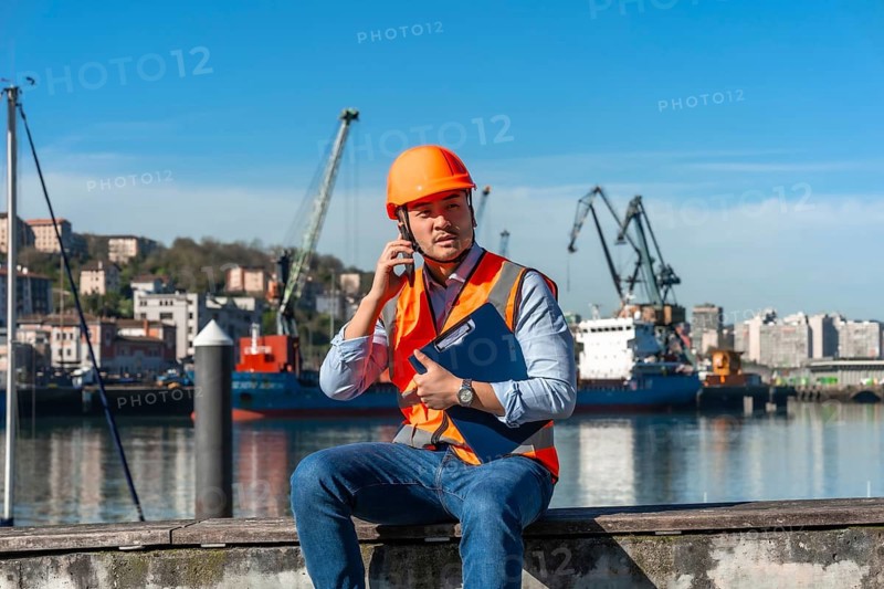 Asian foreman in hard hat and safety vest holds clipboard and talks on smartphone at busy cargo port, overseeing containers, cranes and logistics operations under blue sky