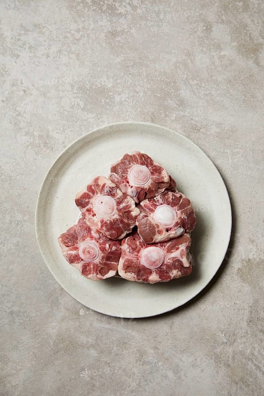 Five raw oxtail pieces are neatly arranged on a round, white ceramic plate. The marbled texture and round vertebrae are visible, showcasing fresh beef cuts perfect for cooking