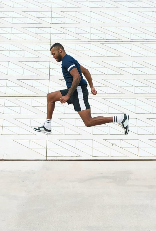 Black athletic man captured mid-air in dynamic action against a modern geometric wall, showcasing power and agility during a high jump training session