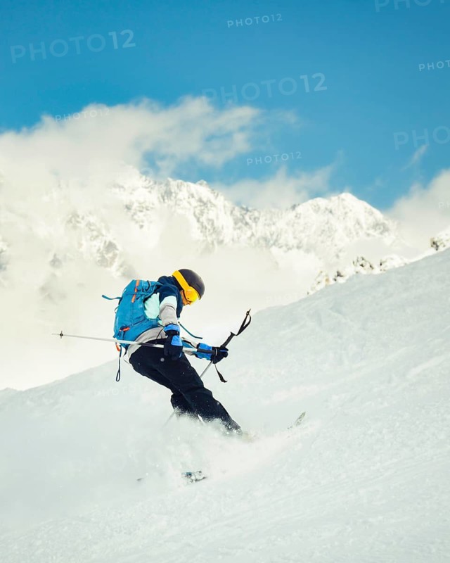 Cool skier performing switch skiing trick backwards on snowy slope with mountains in background