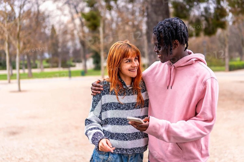 Happy multiethnic couple smiling and enjoying each other's company while using a smartphone in a vibrant park surrounded by nature