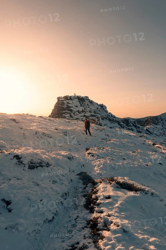 A lone hiker traverses the snow-covered terrains of Siete Picos, Guadarrama Mountains, Madrid. The serene winter landscape is bathed in warm sunset hues