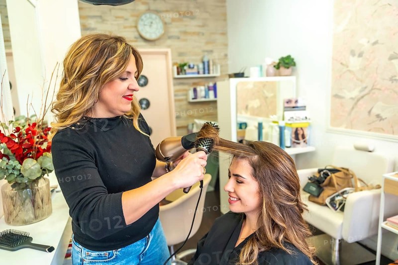 Hairdresser styling a young woman's hair with a blow dryer and round brush, providing professional beauty care service in a bright hair salon environment