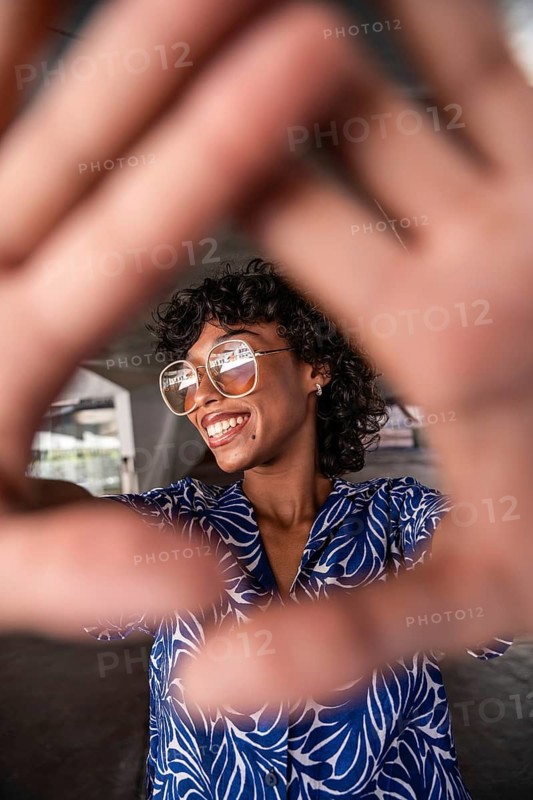 A vibrant woman with curly hair and glasses smiles, framed by her hands in a playful gesture, wearing a blue patterned blouse in an urban setting