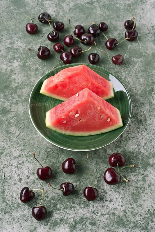 Top view of juicy watermelon slices on a vibrant green plate, surrounded by scattered cherries on a textured green surface. Perfect for summer themed designs