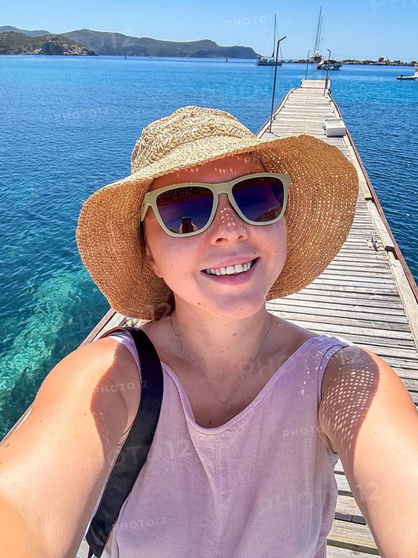 A young Caucasian woman with blonde hair smiles while standing on a wooden pier. She wears a straw hat and sunglasses, with clear blue water in the background