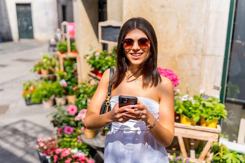 Young woman browsing her smartphone while enjoying a sunny day at a vibrant flower shop, surrounded by colorful blooms and plants