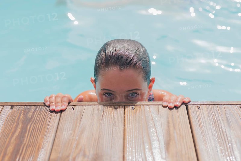 A person peeks over a wooden edge in a calming outdoor pool setting. The clear water reflects sunlight, creating a relaxing and serene atmosphere perfect for leisure