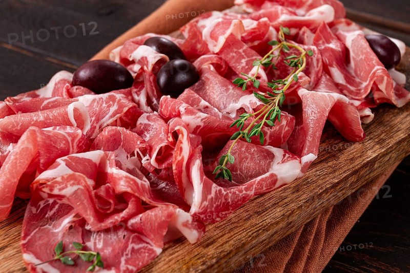 Freshly cut dried meat, served with olives and herbs, on a wooden chopping board, no people