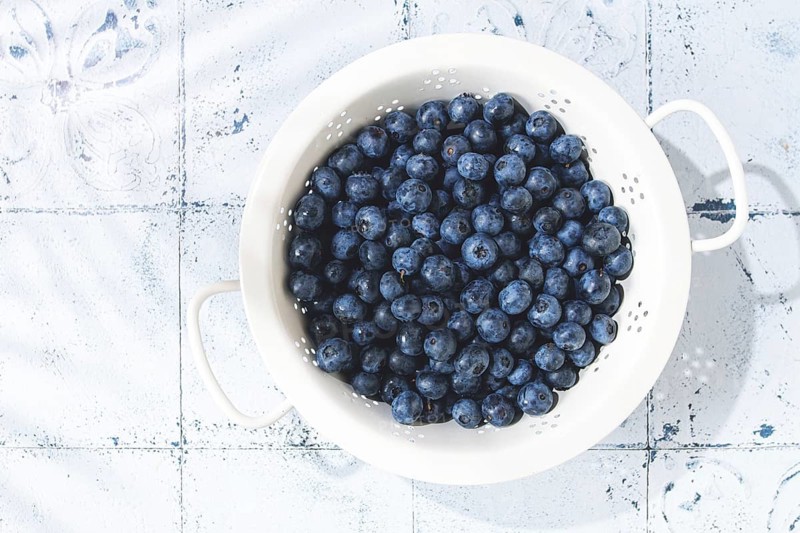 Fresh blueberries sit in a white colander resting on a light blue surface. The colander holds many berries, ready for use in cooking or snacking