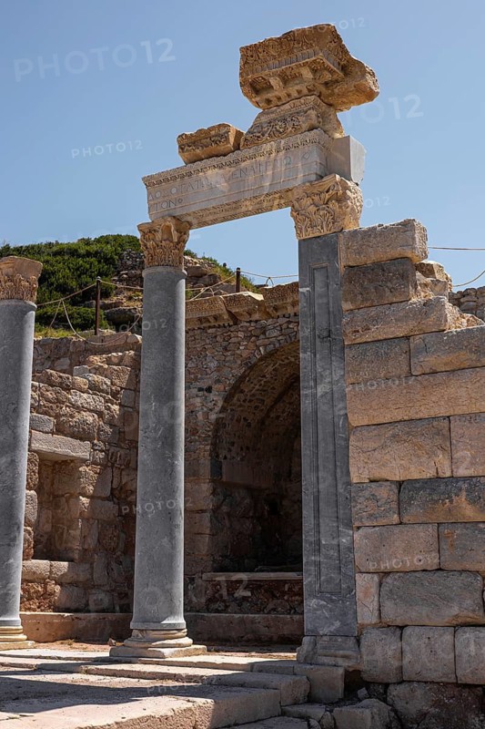 Ancient ruins in Turkiye featuring a stone archway with columns. The structure shows remnants of historical architecture against a clear blue sky