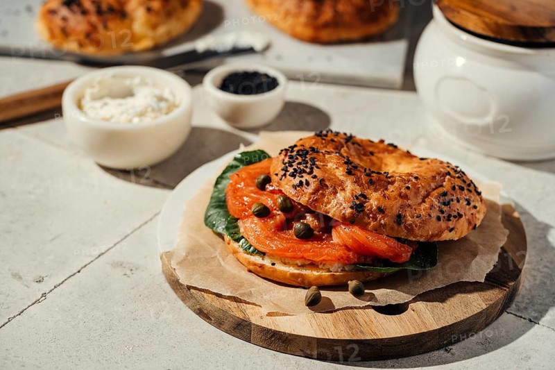 A delicious bagel topped with fresh salmon, capers, and greens, served on a rustic wooden board with cream cheese and spices, creating a perfect breakfast or brunch option