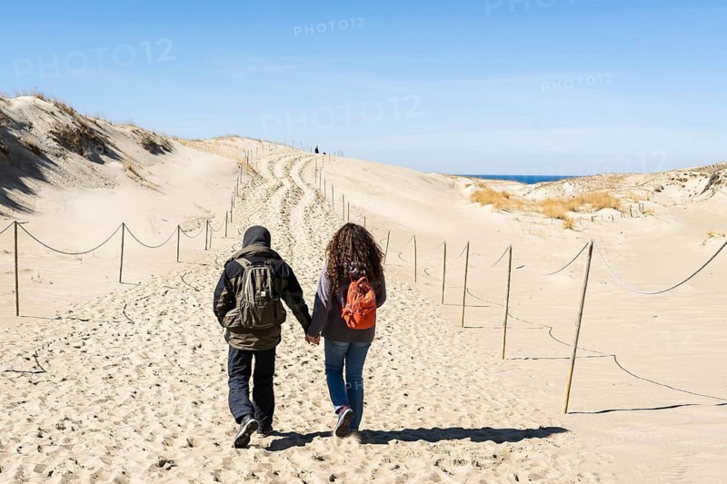 A couple of backpackers hand in hand walk along the hilly sand dunes on the Curonian Spit, Lithuania, A couple walks hand in hand along sandy dunes under a clear blue sky, Curonian Spit, Neringa, Lithuania