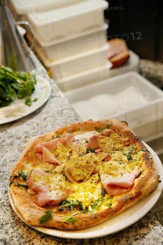 A freshly baked pizza garnished with ham, cheese, and herbs rests on a marble kitchen counter, foregrounding stacked pizza boxes
