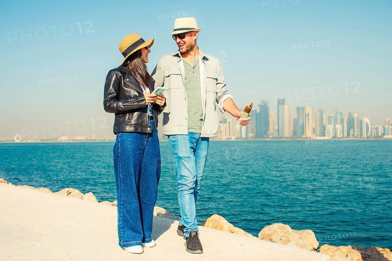 Doha Qatar tourist couple walking Corniche waterfront West Bay with skyline skyscrapers background happy tourists vacation lifestyle Middle East urban city exploration sunny summer fun day bonding