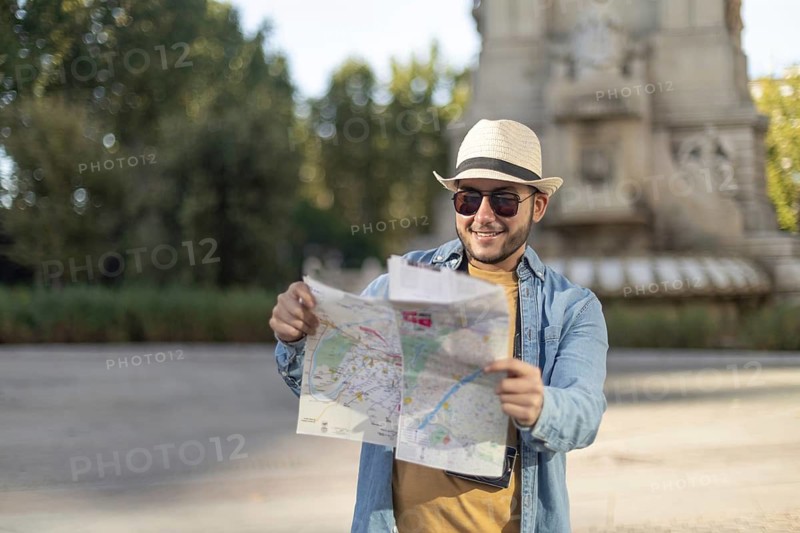 A Latino transgender man wearing a hat and sunglasses enjoys a sunny day out as he explores the city with a map in hand, embodying a spirit of adventure and pride
