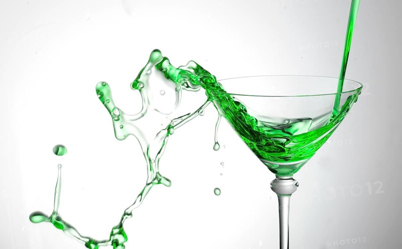 A powerful flood of green liquid pours into a cocktail glass and spills over, studio shot against a white background