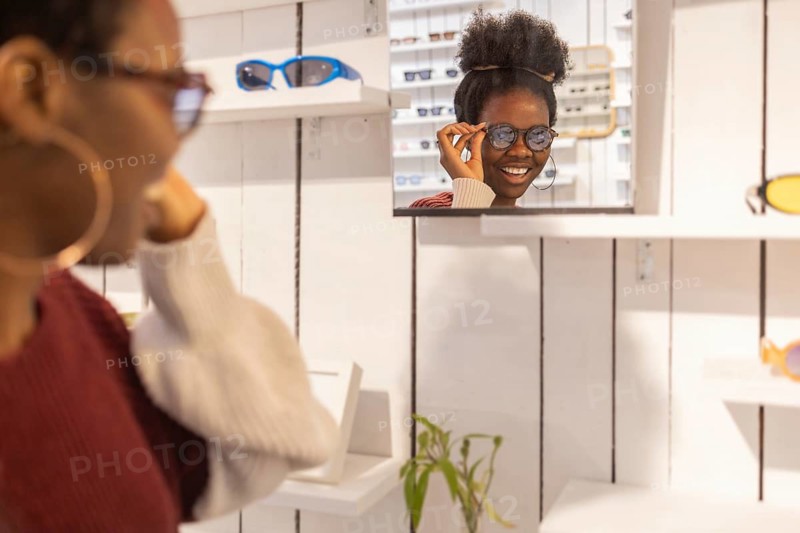 A woman enjoys selecting sunglasses in a trendy store. She admires her choice in the wall mirror, surrounded by various eyewear options in a stylish boutique setting