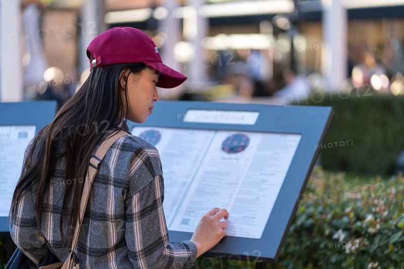 An Asian woman, wearing a red cap and plaid shirt, carefully reads an information panel at a historic site in Avila, Spain, surrounded by lush greenery and a cultural atmosphere