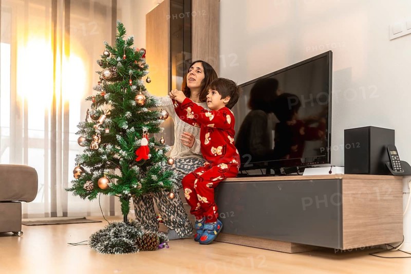 Mother and son dressed in pajamas happily decorating a christmas tree together with ornaments and baubles, celebrating the festive holiday spirit at home
