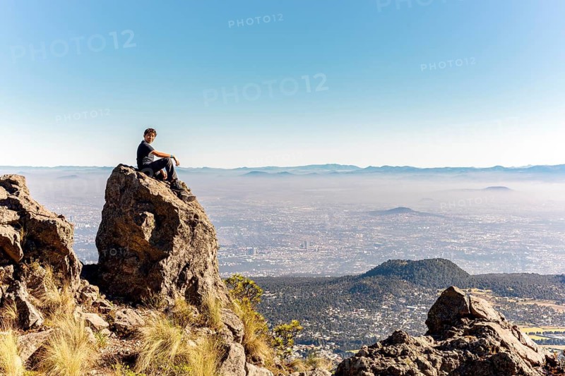 A man sits on a large rock, gazing at a vast cityscape surrounded by mountains The image captures a peaceful moment in nature, offering a stunning view of the horizon