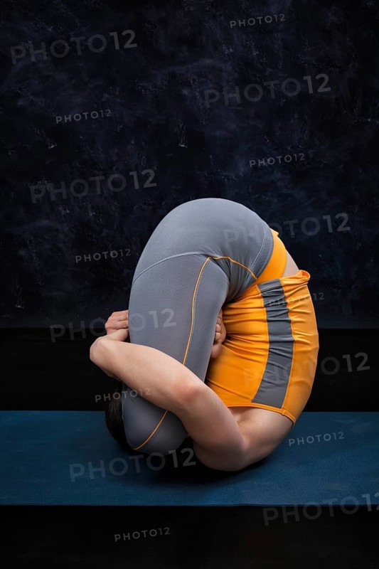 Woman doing Hatha Yoga Ashtanga Vinyasa yoga asana Garbha pindasana, womb embryo pose with lotus on dark grunge background