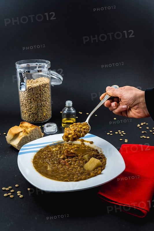 A hand of a young woman eating with a spoon inside a plate of Spanish cooked lentils, along with ingredients