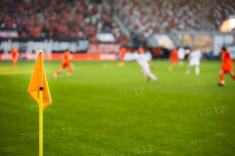 A close-up of a bright yellow corner flag on a grass pitch with a football match and stadium crowd blurred in the background in Lubin, Poland