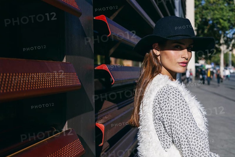 Woman wearing a stylish black hat and white vest, standing against a modern urban backdrop. The sun casts a warm glow, accentuating her chic fashion style