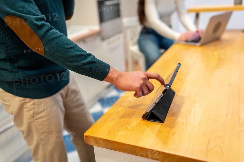 Man using tablet on a wooden kitchen counter while partner works on a laptop in the background, illustrating home multitasking, remote work and everyday digital connectivity