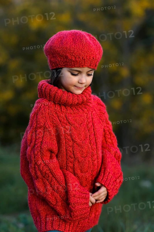 A young girl in a vibrant red woolen hat and sweater is pictured during a serene autumn day, with softly blurred yellow foliage in the background