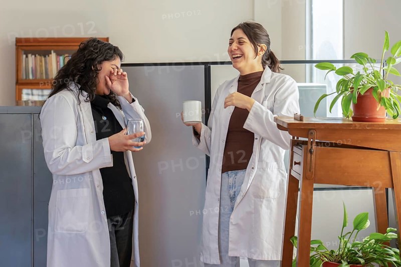 Two young female scientists in white lab coats laughing together during a break, one holding a mug and the other a glass of water, surrounded by indoor plants in a bright office