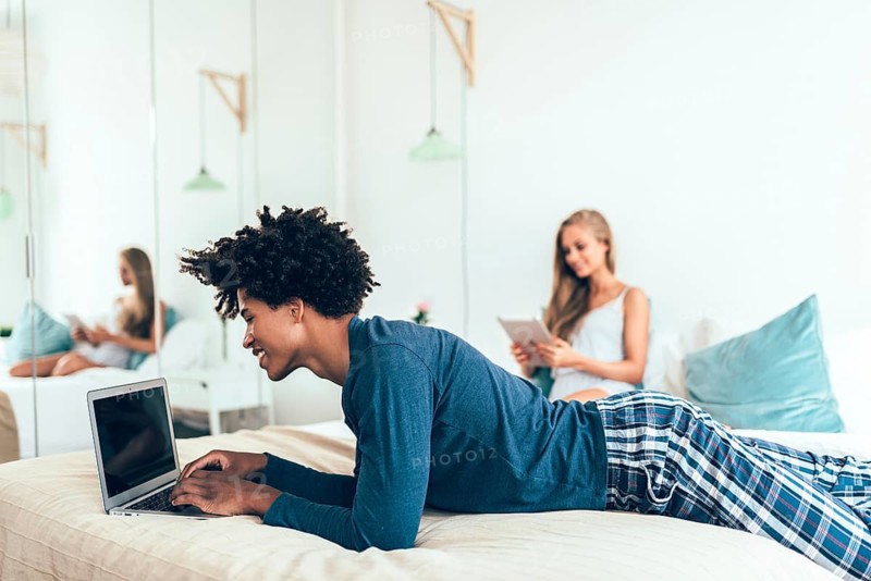 A young man using a laptop and a woman browsing a tablet in a cozy bedroom. They are casually dressed in leisurewear, enjoying a relaxed morning at home