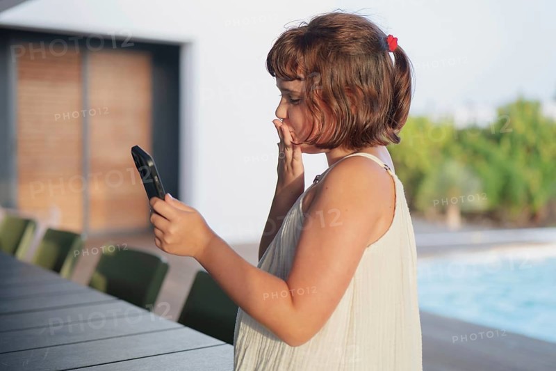 A young girl in a white dress enjoys a sunny summer day by the pool, engaging with her smartphone. Her playful expression captures the essence of a carefree vacation