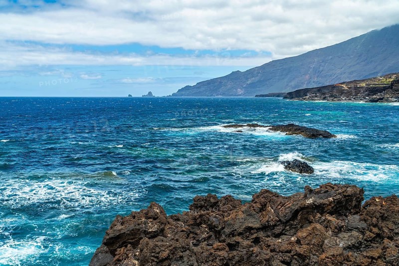 El hierro volcanic coastline featuring dark rocks along the shore, with turquoise atlantic ocean waves breaking and a distant mountain range under a cloudy sky