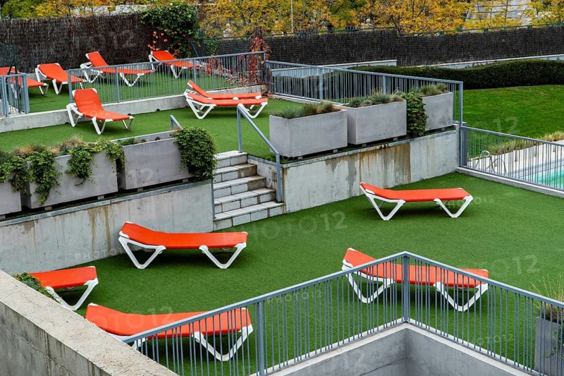 An elevated perspective of a contemporary outdoor pool area featuring bright red-and-white sun loungers scattered across vibrant green artificial turf. The scene includes stylish concrete planters with lush plants