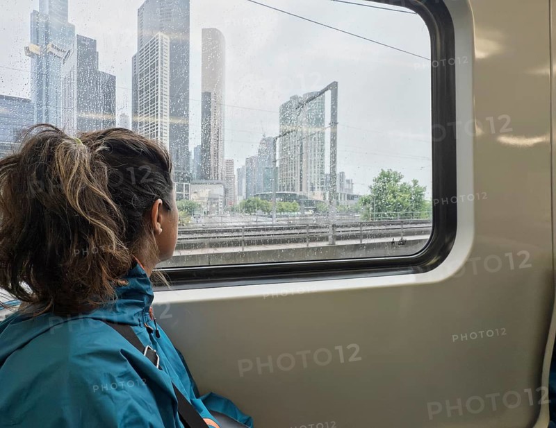 A woman wearing a blue jacket gazes out a rain-speckled train window at the Melbourne skyline. Skyscrapers rise into a cloudy sky, encapsulating urban life