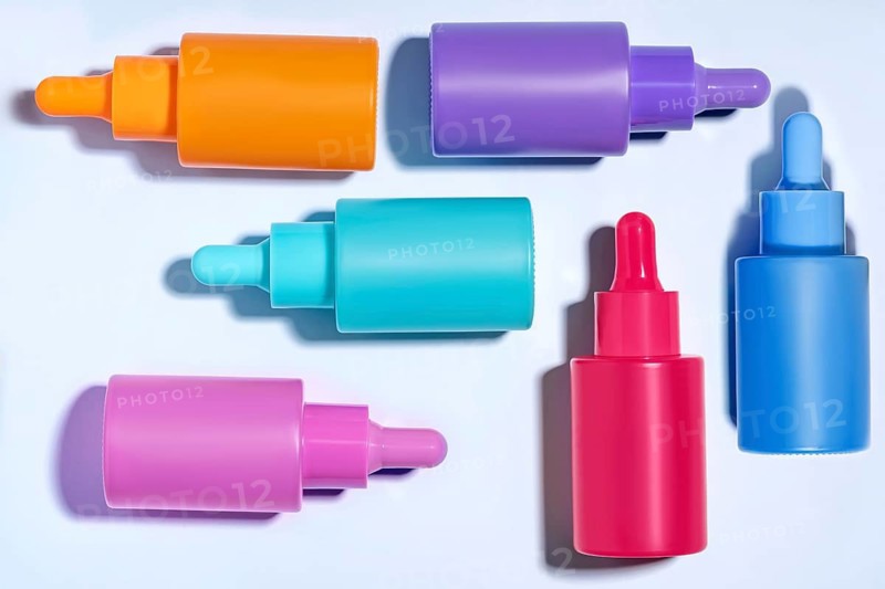 An overhead view of six colorful serum bottles in orange, purple, blue, and pink shades, neatly arranged on a pure white background
