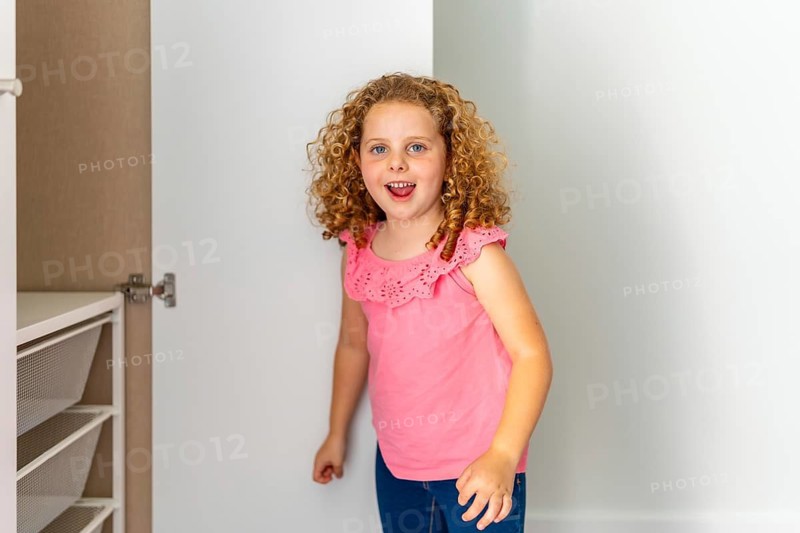 Little girl with curly red hair smiling cheerfully while exploring an empty closet with mesh drawers in her new home, symbolizing the excitement of moving and starting fresh