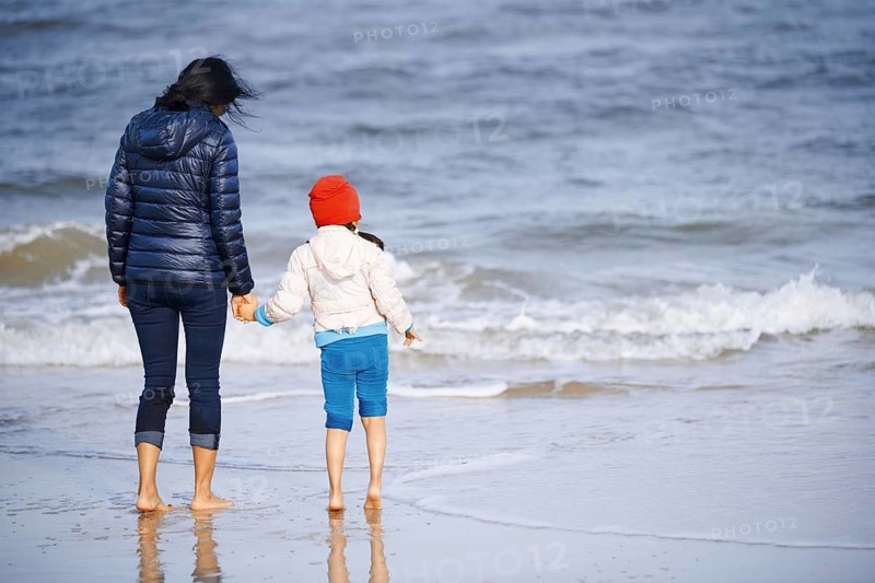 A mother and child, wearing warm clothing, hold hands while walking along a sandy beach in California, United States. The ocean waves gently lap at their feet, encapsulating a serene coastal moment
