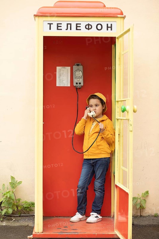 A preteen girl in a yellow jacket using a retro telephone inside a bright red phone booth with Russian text