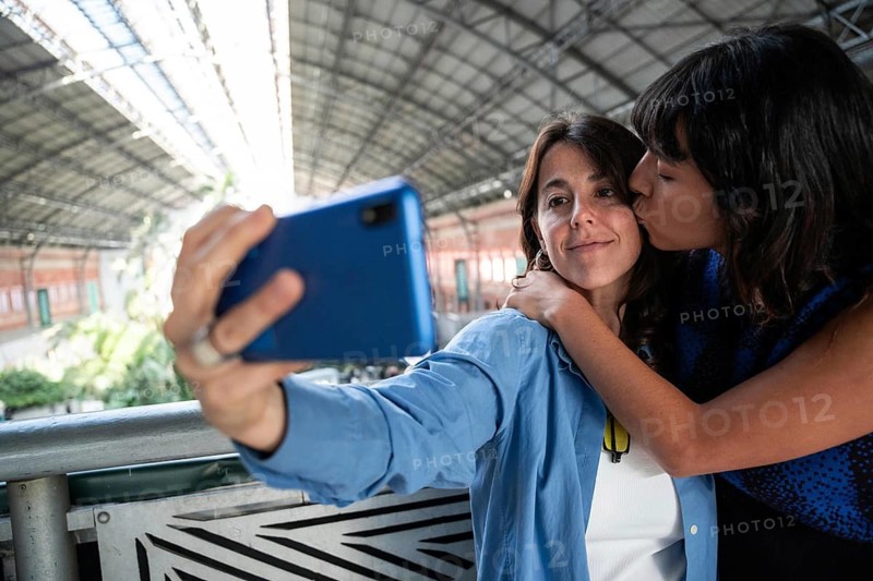 A multiethnic lesbian couple shares a romantic moment at Atocha Station in Madrid. One partner kisses the other's cheek as they take a selfie, capturing their love in the station's beautiful setting