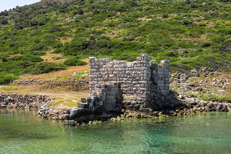 Ruins of an ancient stone structure by the water in Turkiye. Lush green hills surround the site, reflecting a historical landscape