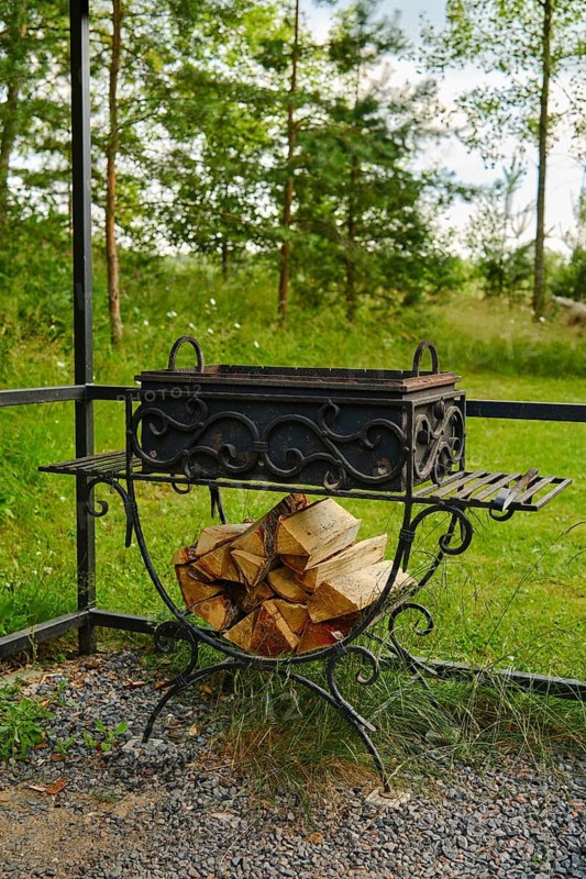 Fire pit stands in garden with neatly stacked firewood beside it. Tall trees and green grass surround backyard. Sunlight filters through the leaves creating a natural atmosphere
