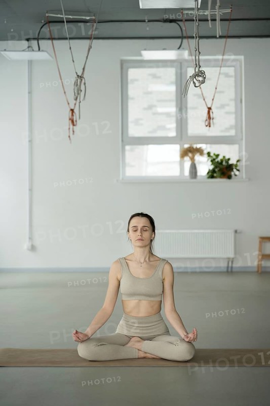 A woman in a yoga studio sits cross legged on a mat, eyes closed, in deep meditation. The serene space includes hanging ropes and soft natural light from a window