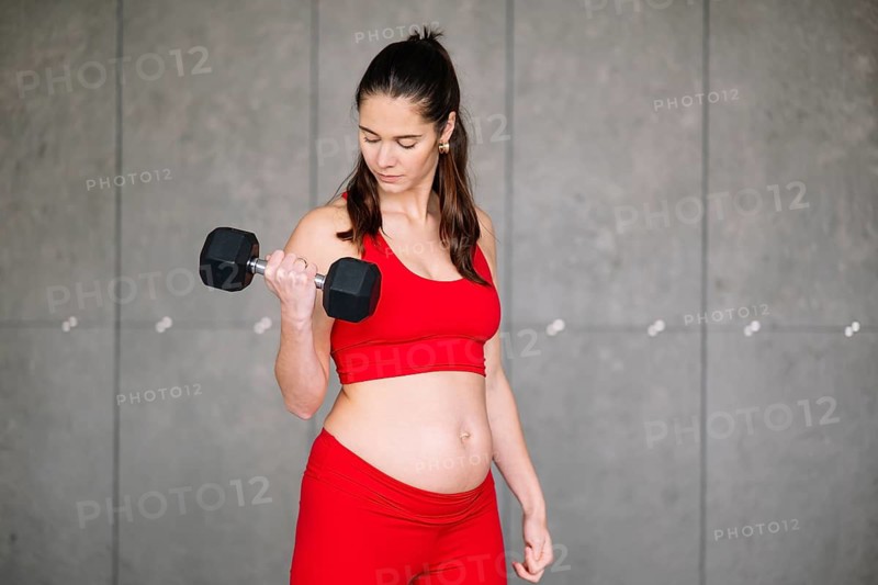 A pregnant woman in a red workout outfit lifts a dumbbell, focusing on strength and wellness during her pregnancy. The background is a neutral gray wall, emphasizing her form